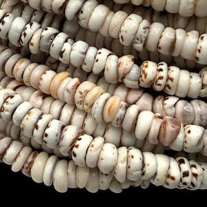 Puede incluir: Una colección de pequeñas cuentas circulares en forma de disco hechas de conchas blancas naturales. Cada cuenta presenta una superficie lisa y pulida con motas marrones irregulares únicas y variaciones naturales en la textura.