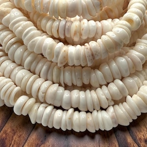 May include: A collection of off-white, circular shell beads strung together in multiple rows. Each bead features a smooth, polished surface with subtle variations in tone and texture, creating a layered, uniform appearance across the strands.