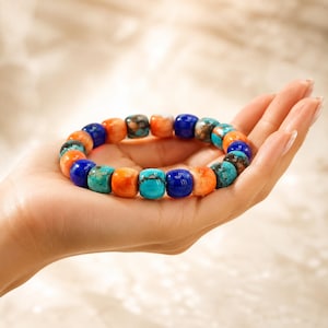 May include: A stretch bracelet composed of smooth, barrel-shaped beads in alternating colours of vibrant orange, deep royal blue, and turquoise with dark brown matrix veining. The beads are arranged in a repeating pattern around the circular band.