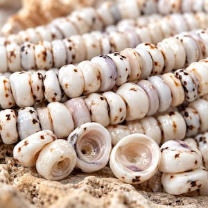 May include: A collection of small, round, disc-shaped beads crafted from natural white shells. Each bead features a smooth, polished surface with irregular brown speckles and subtle purple-toned centers, arranged in multiple parallel rows.