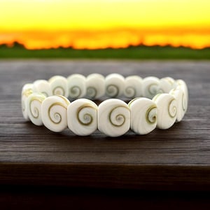 May include: A white shell bracelet featuring a spiral pattern. The bracelet is crafted from natural shell beads and is a beautiful and unique piece of jewellery.