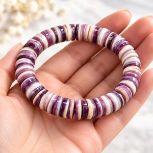 May include: A stretch bracelet composed of numerous small, flat, circular disc beads. The beads feature a repeating color pattern of deep purple, light lavender, and creamy white, creating a smooth, polished, and uniform appearance throughout.