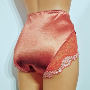 May include: Peach-colored high-waisted underwear made of a shiny satin material. The side panels feature a delicate floral lace design with a scalloped edge. The underwear has a smooth, seamless appearance.
