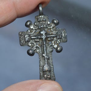 May include: A silver-toned, ornate cross pendant with a figure of Jesus Christ on the front. The cross has a decorative design with small balls at the ends of each arm.