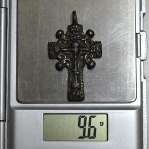 May include: A silver-toned cross pendant with intricate details and a weight of 9.6 grams. The cross is on a digital scale.