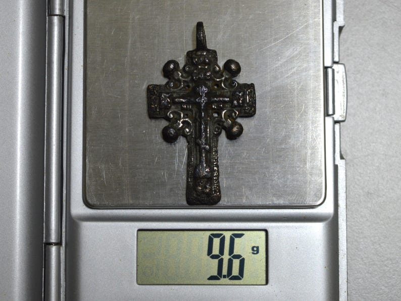 May include: A silver-toned cross pendant with intricate details and a weight of 9.6 grams. The cross is on a digital scale.