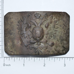 Könnte beinhalten: Eine Nahaufnahme einer angelaufenen, bronzenen Schnalle mit einem erhabenen, detaillierten Bild des russischen Kaiseradlers. Der Adler hält ein Zepter und eine Kugel und trägt eine Krone auf dem Kopf. Die Schnalle ist etwa 5 cm breit.