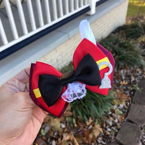 Puede incluir: Un lazo rojo, negro y amarillo con un adorno de encaje blanco y un dije de llave plateado. El lazo está decorado con una pluma blanca.