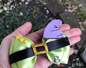 May include: A green satin bow with a black belt and gold buckle. There are three yellow buttons on the bow and three clear rhinestones on the other side. A purple felt hat with a black line is attached to the bow.