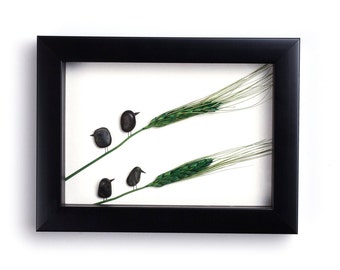 May include: Framed artwork featuring a minimalist design. Black framed art with a white background. The artwork depicts four small, dark birds perched on green wheat stalks. The birds are made of small, smooth stones.