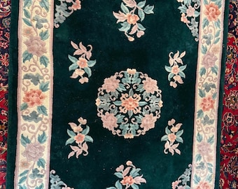 Op de afbeelding: Een rechthoekig, donkergroen tapijt met een bloemenontwerp. Het tapijt heeft een centraal medaillon en bloemmotieven in de hoeken in tinten roze, perzik en blauw. De rand is crèmekleurig met bloempatronen. Het tapijt heeft een franje.