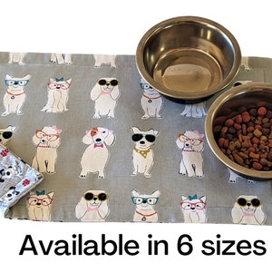 May include: A gray and white dog-themed pet food mat with two stainless steel bowls. The mat features a repeating pattern of dogs wearing sunglasses. The text "Available in 6 sizes" is printed below the mat.