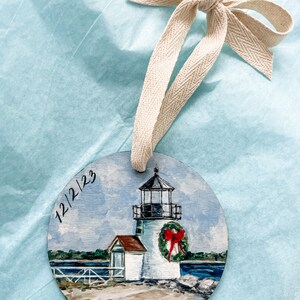May include: A hand-painted wooden bauble depicting a white lighthouse with a red bow on a wreath. The lighthouse is situated on a small island with a sandy beach and blue water. The bauble is dated "12/2/23".