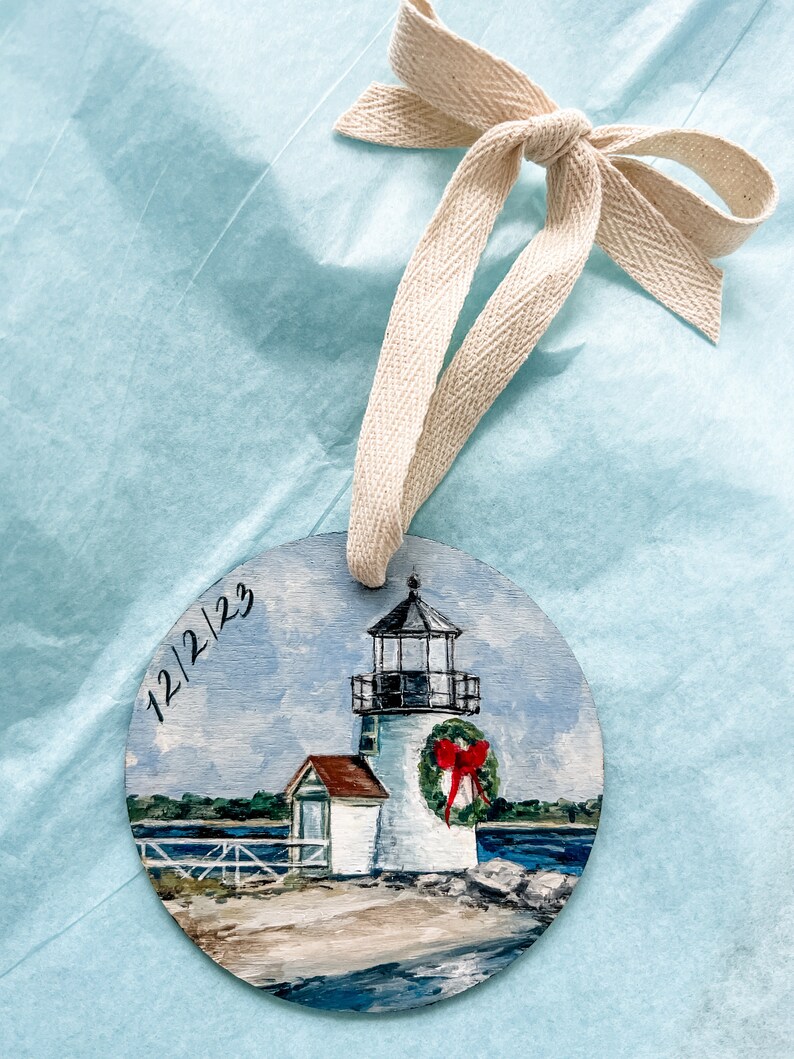 May include: A hand-painted wooden bauble depicting a white lighthouse with a red bow on a wreath. The lighthouse is situated on a small island with a sandy beach and blue water. The bauble is dated "12/2/23".