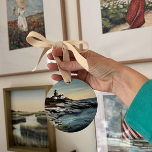 May include: A round wooden bauble painted with a seascape scene featuring a lighthouse on a rocky shore. The bauble is tied with a cream-coloured ribbon.