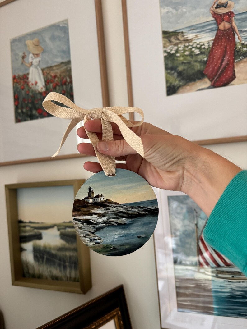 May include: A round wooden bauble painted with a seascape scene featuring a lighthouse on a rocky shore. The bauble is tied with a cream-coloured ribbon.