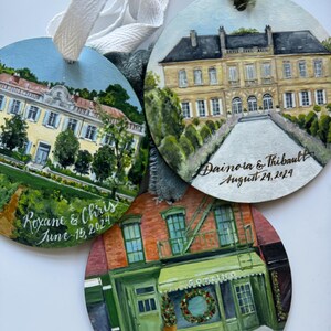 May include: Three round wooden ornaments with painted scenes of buildings. The top ornament features a yellow building with a green roof and white trim. The text "Dainora & Thibault August 24, 2024" is written in gold script. The middle ornament features a red brick building with a green awning and the text "GOTTIINO" above the door. The text "Engaged March 30, 2024" is written in gold script. The bottom ornament features a yellow building with a green roof and white trim. The text "Roxane & Chris June 15, 2024" is written in gold script.