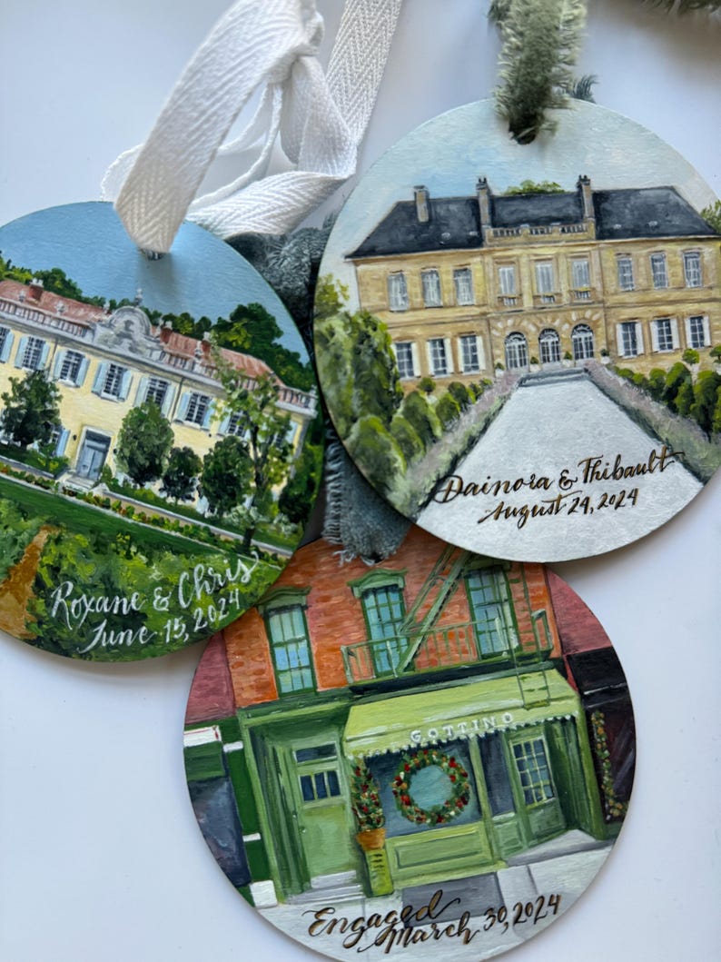 May include: Three round wooden ornaments with painted scenes of buildings. The top ornament features a yellow building with a green roof and white trim. The text "Dainora & Thibault August 24, 2024" is written in gold script. The middle ornament features a red brick building with a green awning and the text "GOTTIINO" above the door. The text "Engaged March 30, 2024" is written in gold script. The bottom ornament features a yellow building with a green roof and white trim. The text "Roxane & Chris June 15, 2024" is written in gold script.