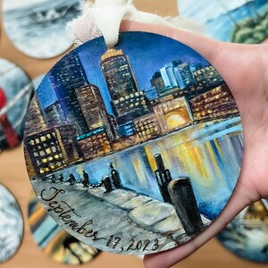 May include: A round wooden ornament featuring a painted cityscape scene. The city is depicted at night with a reflection of the city lights in the water. The ornament is decorated with the date "September 17, 2023" in cursive.