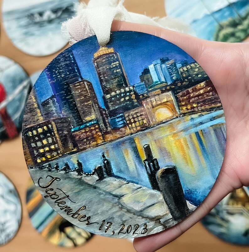 May include: A round wooden ornament featuring a painted cityscape scene. The city is depicted at night with a reflection of the city lights in the water. The ornament is decorated with the date "September 17, 2023" in cursive.