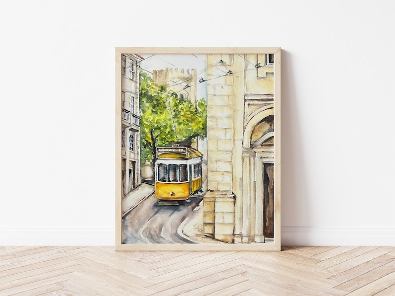 May include: A watercolour painting of a yellow tram travelling down a narrow street in a European city. The tram is surrounded by buildings with stone walls and arched doorways. The painting is framed in a light wood frame.
