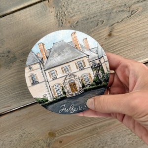May include: A hand-painted round wooden ornament depicting a detailed white house with a grey roof, brown doors, and green landscaping. The date "July 6, 20" is written in black cursive on the bottom.