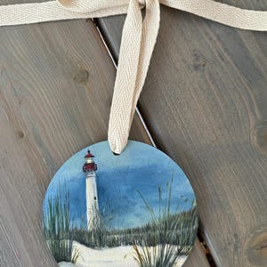 May include: A round wooden ornament painted with a lighthouse on a beach with the date September 6, 2024 written below. The ornament is tied with a beige ribbon.