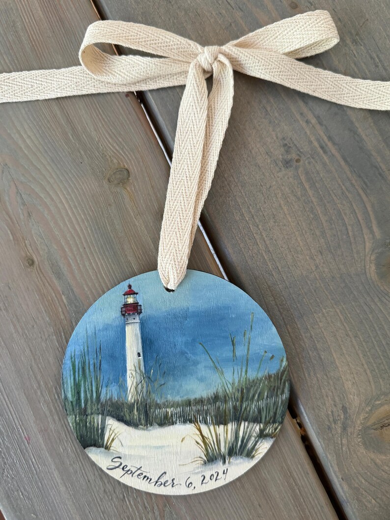 May include: A round wooden ornament painted with a lighthouse on a beach with the date September 6, 2024 written below. The ornament is tied with a beige ribbon.