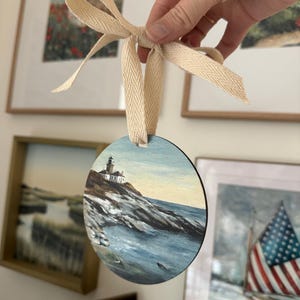 May include: A round wooden bauble featuring a painted scene of a lighthouse on a rocky coastline with blue water. The bauble is tied with a white ribbon.