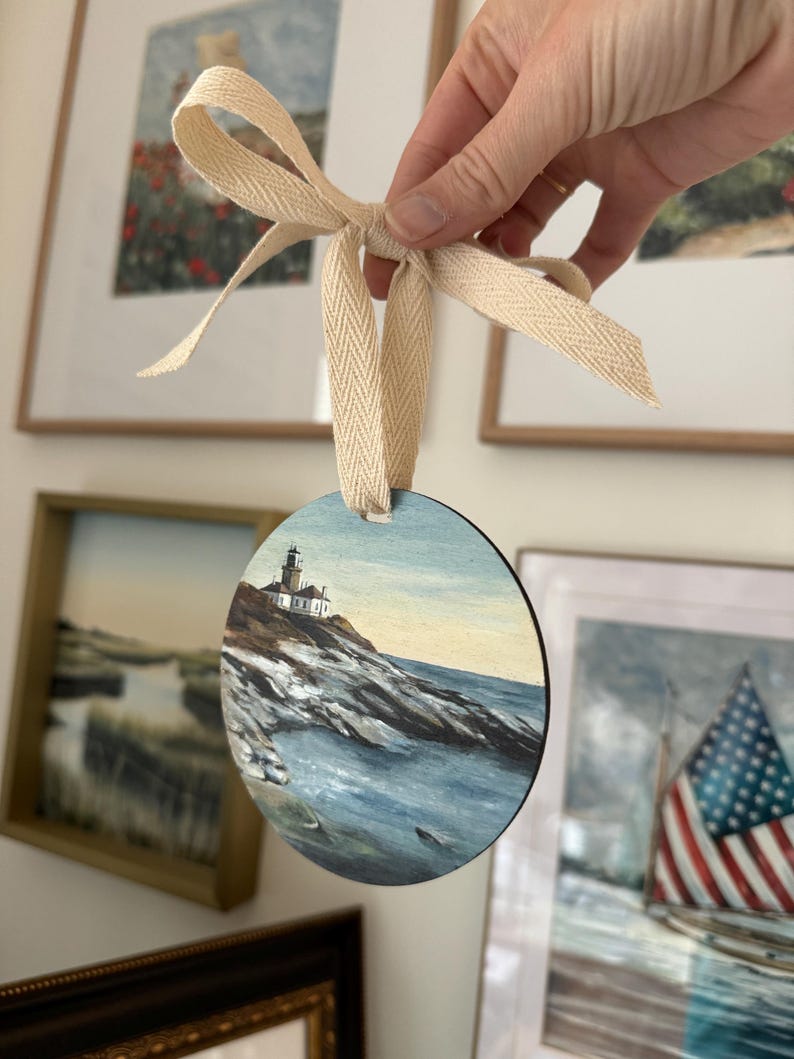 May include: A round wooden bauble featuring a painted scene of a lighthouse on a rocky coastline with blue water. The bauble is tied with a white ribbon.