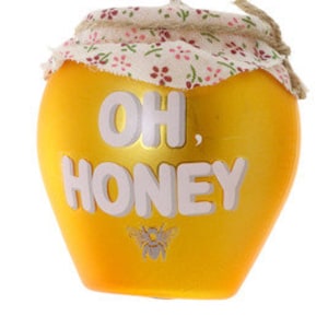 Puede incluir: Un adorno de tarro de miel amarillo con una tapa de tela blanca y el texto "Oh, Honey" en letras blancas. Una pequeña ilustración de abeja está debajo del texto.