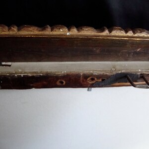 Puede incluir: Primer plano de una secci&oacute;n de un marco de fotos de madera vintage. El marco tiene un acabado marr&oacute;n oscuro con detalles dorados ornamentados en el borde superior. Un soporte met&aacute;lico para colgar est&aacute; fijado en la parte posterior. El marco est&aacute; montado en una pared blanca.
