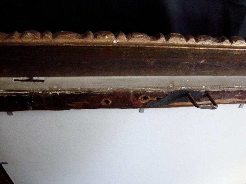 Puede incluir: Primer plano de una secci&oacute;n de un marco de fotos de madera vintage. El marco tiene un acabado marr&oacute;n oscuro con detalles dorados ornamentados en el borde superior. Un soporte met&aacute;lico para colgar est&aacute; fijado en la parte posterior. El marco est&aacute; montado en una pared blanca.