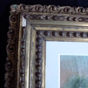 Puede incluir: Primer plano de una secci&oacute;n de un cuadro enmarcado. El marco dorado y ornamentado presenta intrincadas tallas y un borde de cuentas. La pintura en su interior tiene un borde blanco y un atisbo de un paisaje.