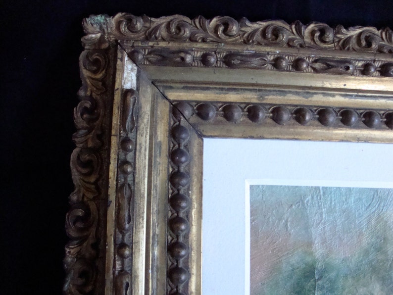 Puede incluir: Primer plano de una secci&oacute;n de un cuadro enmarcado. El marco dorado y ornamentado presenta intrincadas tallas y un borde de cuentas. La pintura en su interior tiene un borde blanco y un atisbo de un paisaje.