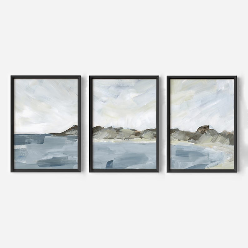 May include: Set of three framed abstract landscape paintings. Each piece features a coastal scene with blue water, brown hills, and a cloudy sky. The artwork is in a modern style with a minimalist aesthetic.