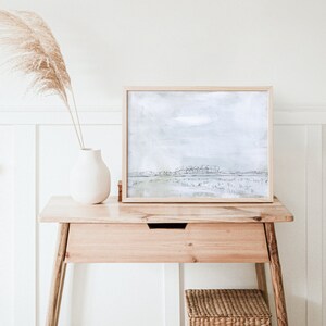 May include: A framed abstract landscape painting with a light blue and green color scheme. The painting depicts a field with a line of trees in the distance. The frame is a light wood color.