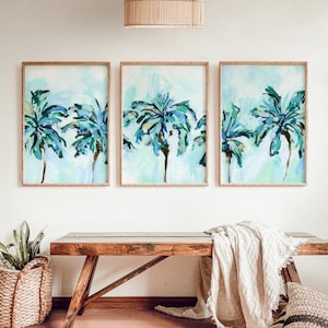 May include: Three framed abstract paintings of palm trees in shades of blue and green on a white wall above a wooden bench with a white throw blanket.