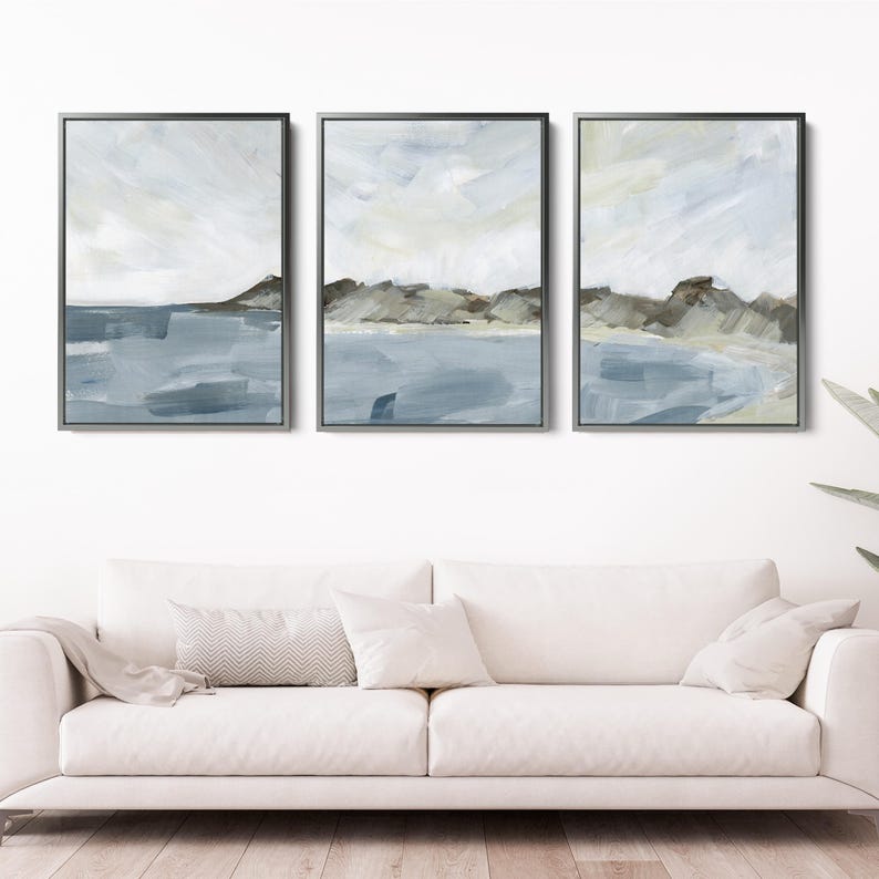 May include: Set of three framed abstract landscape paintings. Each piece features a coastal scene with a muted color palette of blues, grays, and browns. The artwork is displayed above a white sofa, adding a touch of modern art to the room.
