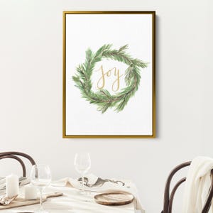 May include: A framed print featuring a watercolor wreath of green pine needles. The word "Joy" is written in gold script within the wreath. The frame is gold, and the background is white.