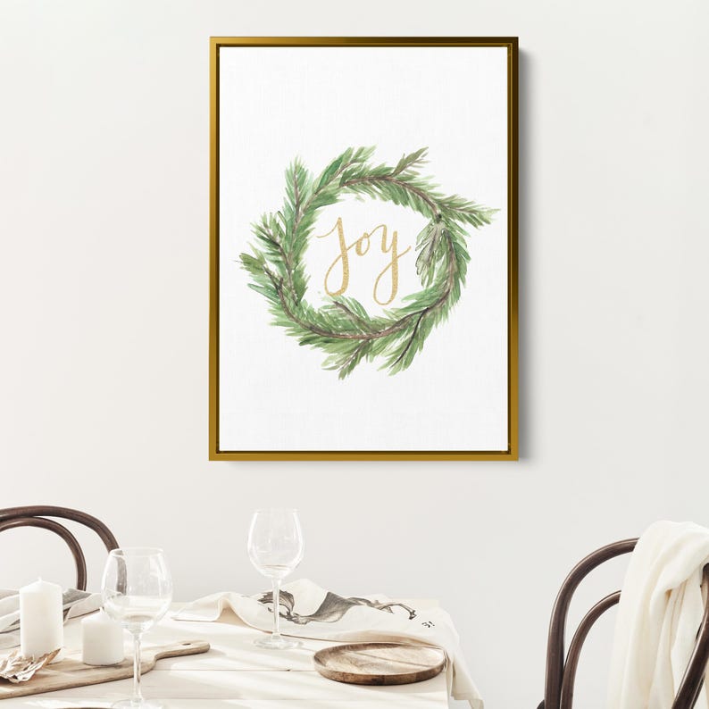 May include: A framed print featuring a watercolor wreath of green pine needles. The word "Joy" is written in gold script within the wreath. The frame is gold, and the background is white.