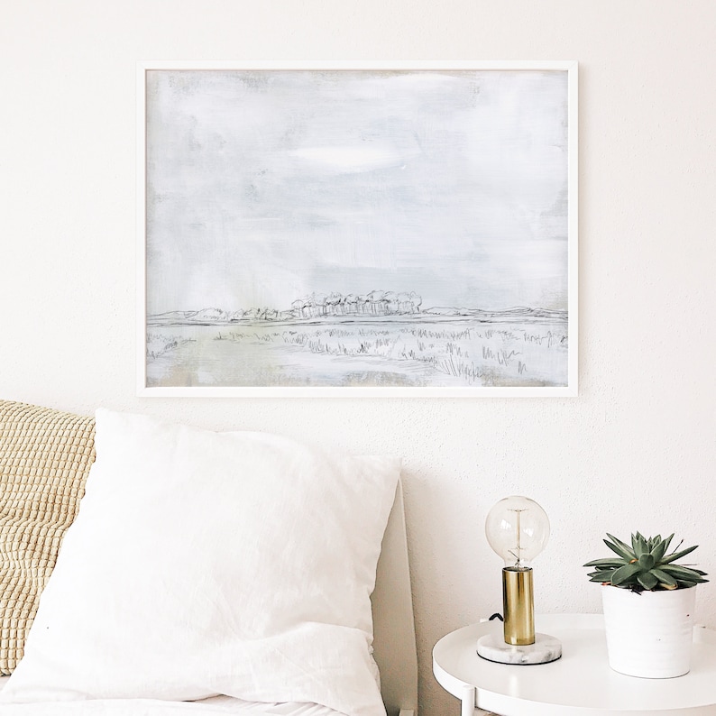 May include: A framed black and white watercolor painting of a landscape with a row of trees in the distance. The sky is a light blue with a hint of gray. The painting is on a white wall above a white bed with a tan pillow.