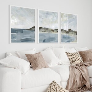 May include: Three framed abstract landscape paintings in shades of blue, gray, and brown. The artwork depicts a coastal scene with brushstrokes suggesting water, sky, and land. The paintings are displayed above a white sofa with pillows and a throw.