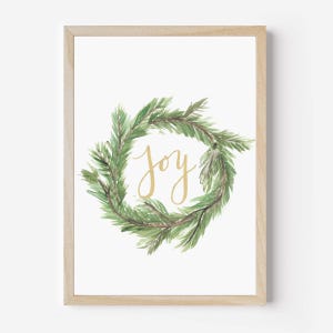 May include: A framed print featuring a watercolor wreath of green pine needles. The word "joy" is written in gold script in the center of the wreath. The frame is made of light-colored wood.