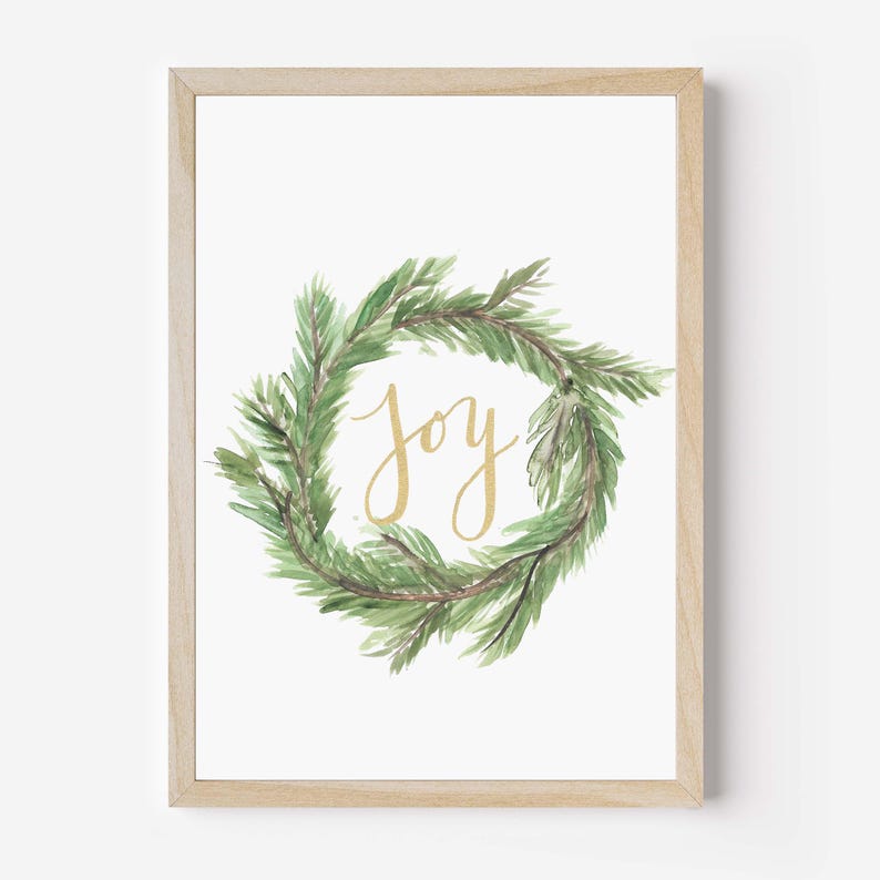 May include: A framed print featuring a watercolor wreath of green pine needles. The word "joy" is written in gold script in the center of the wreath. The frame is made of light-colored wood.