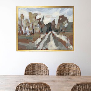 May include: An abstract landscape painting in a gold frame. The artwork depicts a road leading into a forest with trees in shades of brown, beige, and green. The sky is a mix of white and light blue. The painting is displayed above a wooden table and wicker chairs.