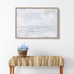 May include: A framed black and white sketch of a seascape with waves crashing on the shore. The background is a light gray with a hint of blue. The frame is a light wood color. A small basket of blue flowers sits on a fringed bench in front of the artwork.