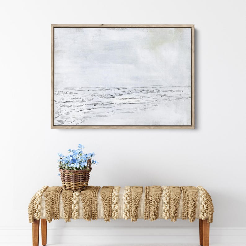 May include: A framed black and white sketch of a seascape with waves crashing on the shore. The background is a light gray with a hint of blue. The frame is a light wood color. A small basket of blue flowers sits on a fringed bench in front of the artwork.