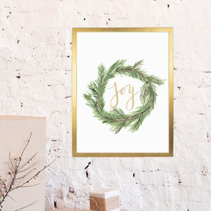 May include: A framed print featuring a watercolor wreath of green pine needles. The word "Joy" is written in gold script within the wreath. The frame is gold, and the background is white.