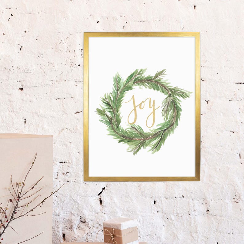 May include: A framed print featuring a watercolor wreath of green pine needles. The word "Joy" is written in gold script within the wreath. The frame is gold, and the background is white.